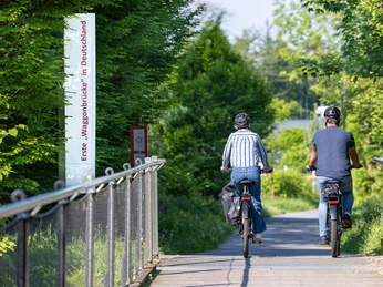 Tour à vélo sur le pont Waggong à Heiligenhaus Deux personnes traversent un pont à vélo, entourées de végétation verte et de soleil.