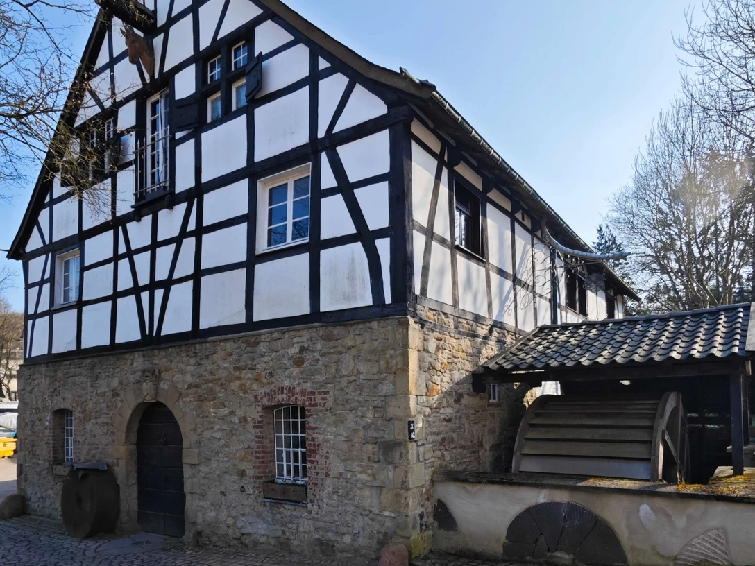 Rindersberger Mill in Essen Half-timbered house made of stone and wood with a water wheel, surrounded by trees, under a sunny sky.