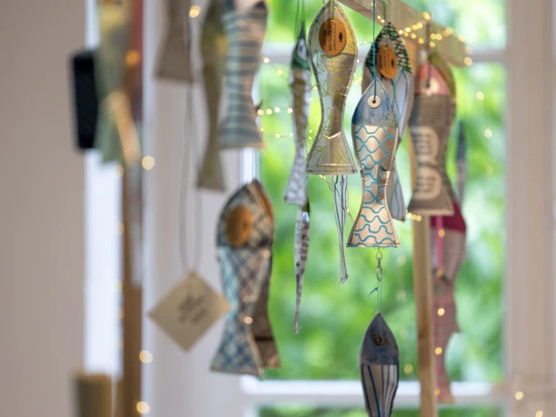 Papiermarkt 2024 Dekorative Papierfische und Lichterketten hängen an einem Holzgestell vor einem Fenster mit grünem Ausblick.