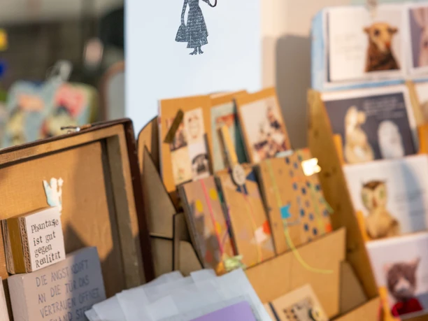 Papiermarkt 2024 Ein bunter Flohmarktstand mit handgefertigten Kunstwerken, Karten und einem hängenden Regenschirm-Mobilé.