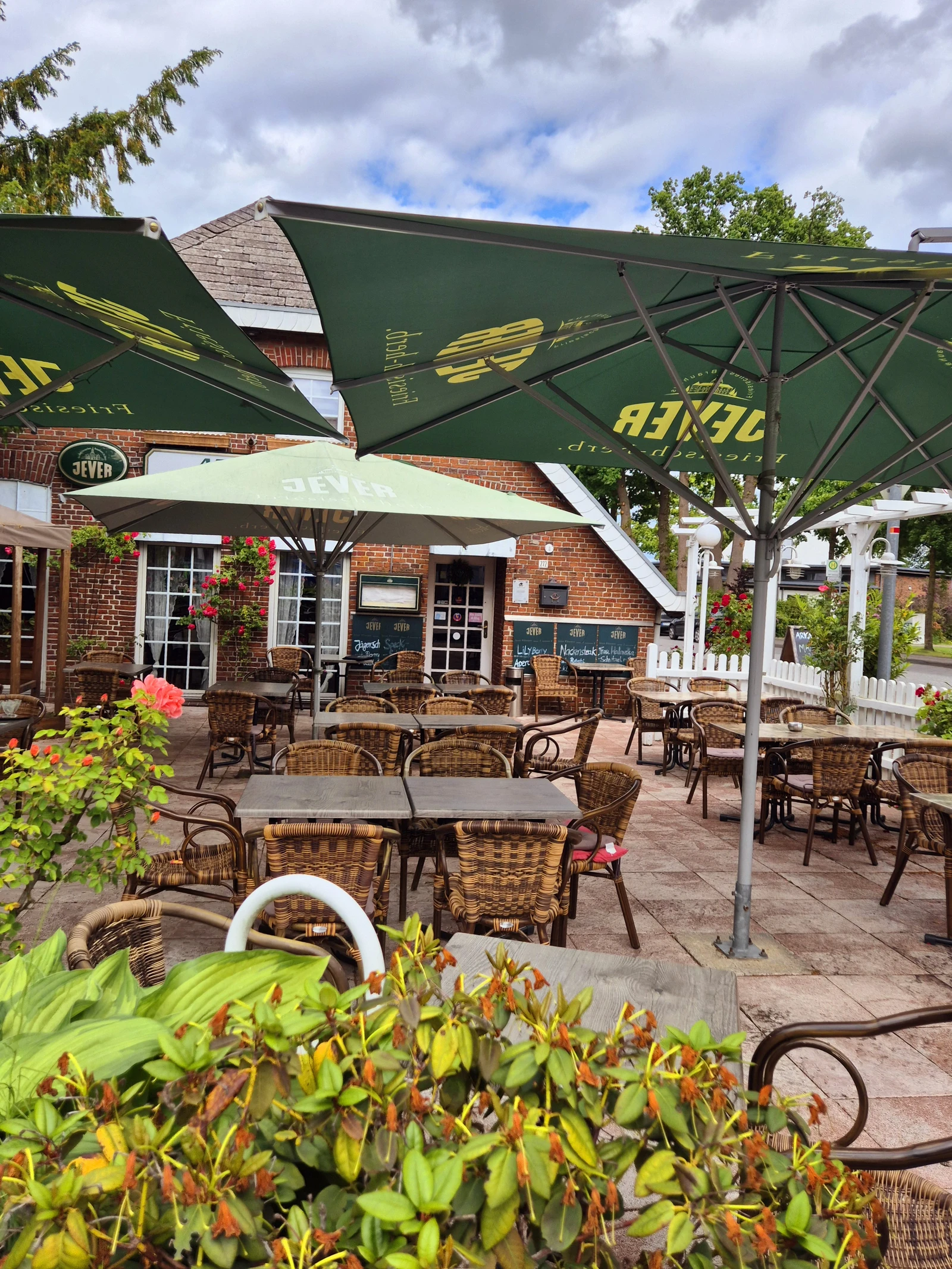 Gemütlicher Biergarten mit grünen Sonnenschirmen und Korbmöbeln, umgeben von Blumen und Bäumen.