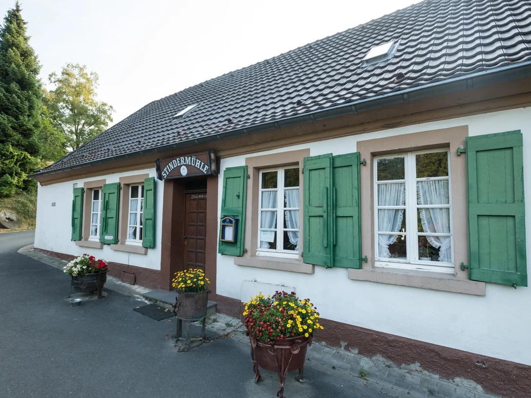 Stindermühle w Erkrath.jpg Biały budynek z zielonymi okiennicami i szyldem z napisem „Stindermühle”, otoczony doniczkami z kwiatami.