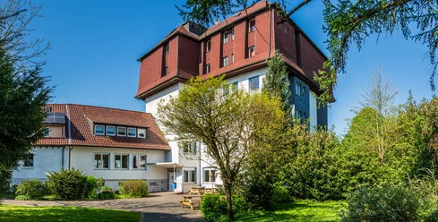 Multi-story building with a red roof and white walls, surrounded by green trees and lawns.