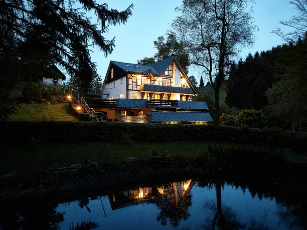 Buitenaanzicht bij schemering Een idyllisch huis aan de rand van het bos wordt verlicht door de schemering en weerspiegeld in de rustige vijver.