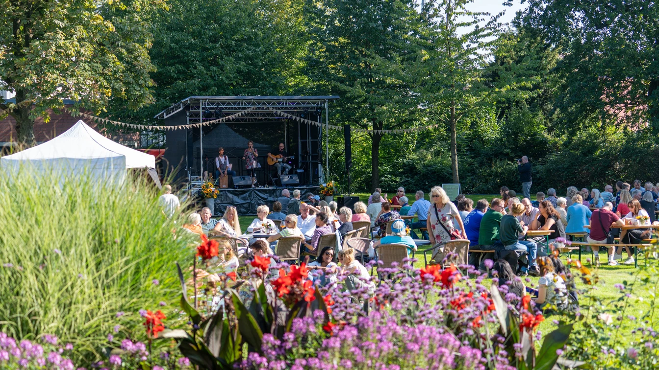 Open-air concert in the countryside with musicians on stage and audience at tables in the sunshine.