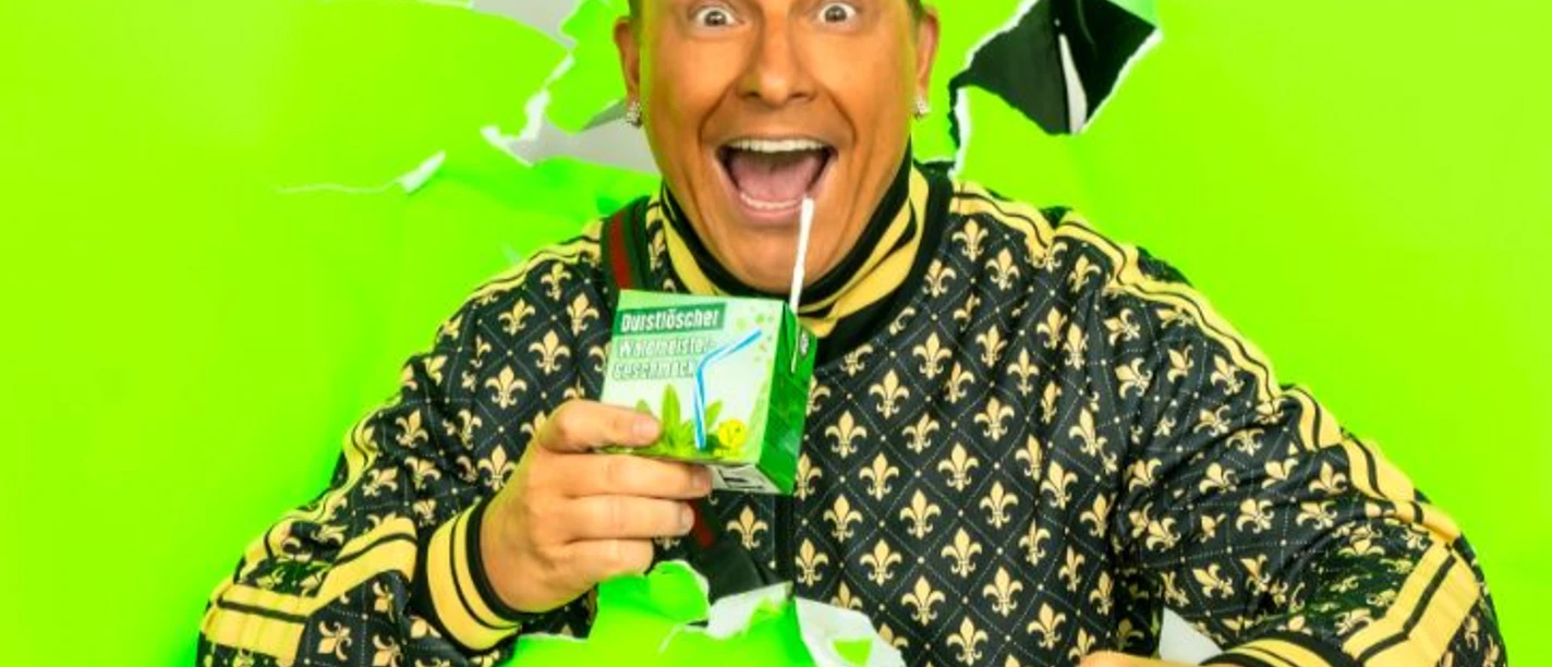 Dennis da Hürth Un uomo allegro, vestito con abiti vistosi, beve da un cartone di bevande su uno sfondo verde chiaro.