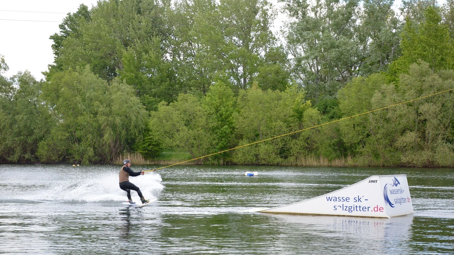 Person fährt Wakeboard am Seilzug über den Salzgittersee vor grünem Ufer mit Bäumen.