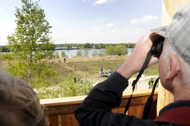 Vogelkijktoren op Bargerveen in Twist ©Naturpark Moor-Veenland (29).jpg Een man observeert de mensen en het landschap bij het meer in het natuurpark Moor-Veenland vanuit een houten uitkijktoren met een verrekijker.