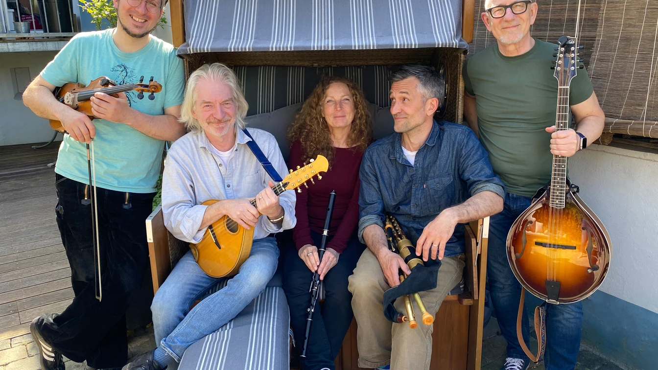 Fünf Personen mit Musikinstrumenten sitzen und stehen um eine Strandkorbbank auf gepflastertem Boden.