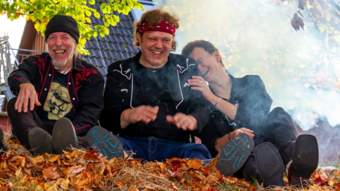 Drei Personen sitzen auf einem mit Herbstlaub bedeckten Boden vor einem Hausdach, umgeben von Rauch und Bäumen mit grünen und gelben Blättern