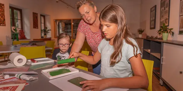 Zwei Mädchen basteln im Geoportal Porphyrhaus unter Anleitung. Sie sitzen an einem Tisch und machen Linoldruck.