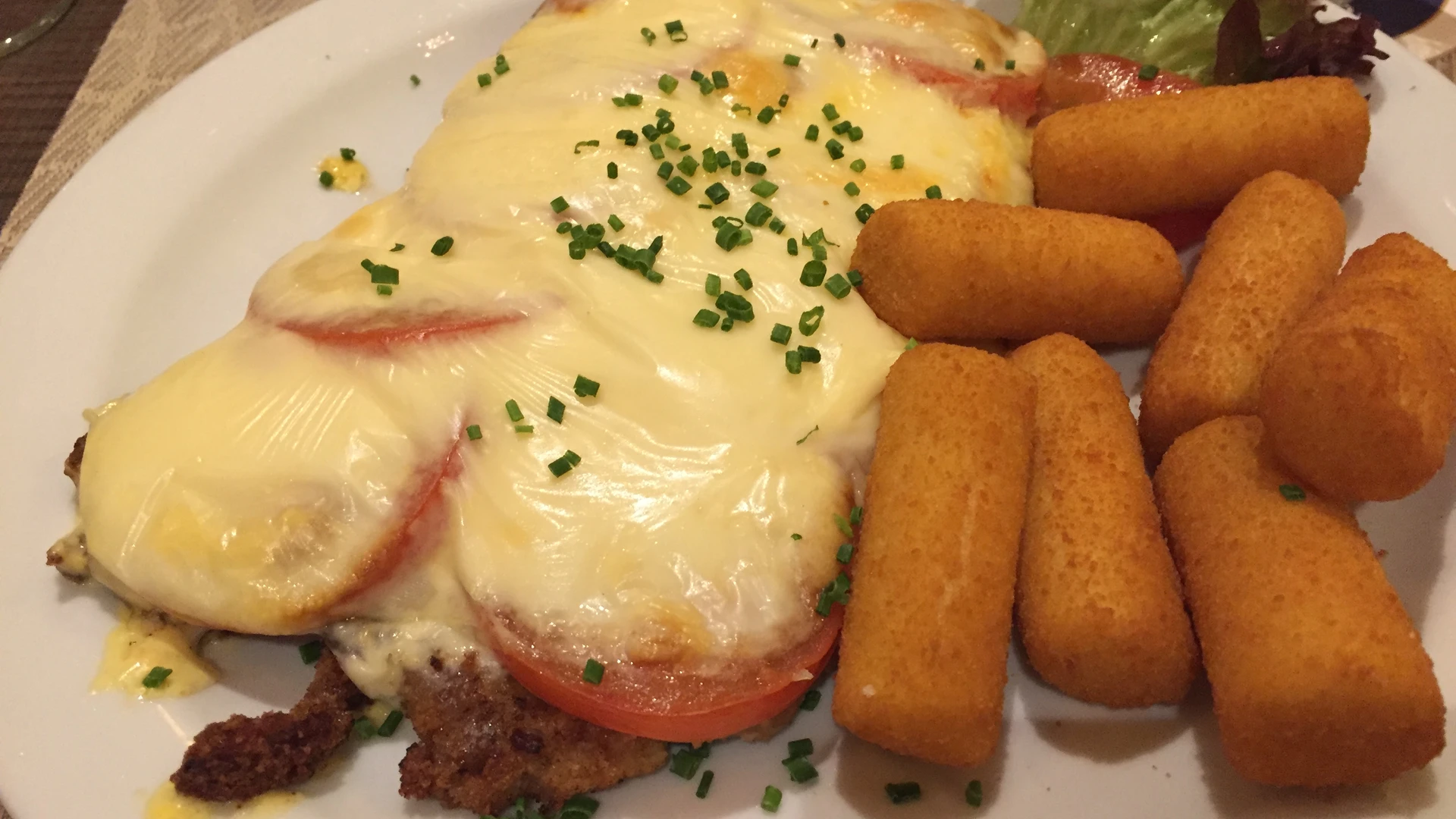 An oven-baked schnitzel with tomatoes, melted cheese and croquettes on a plate.