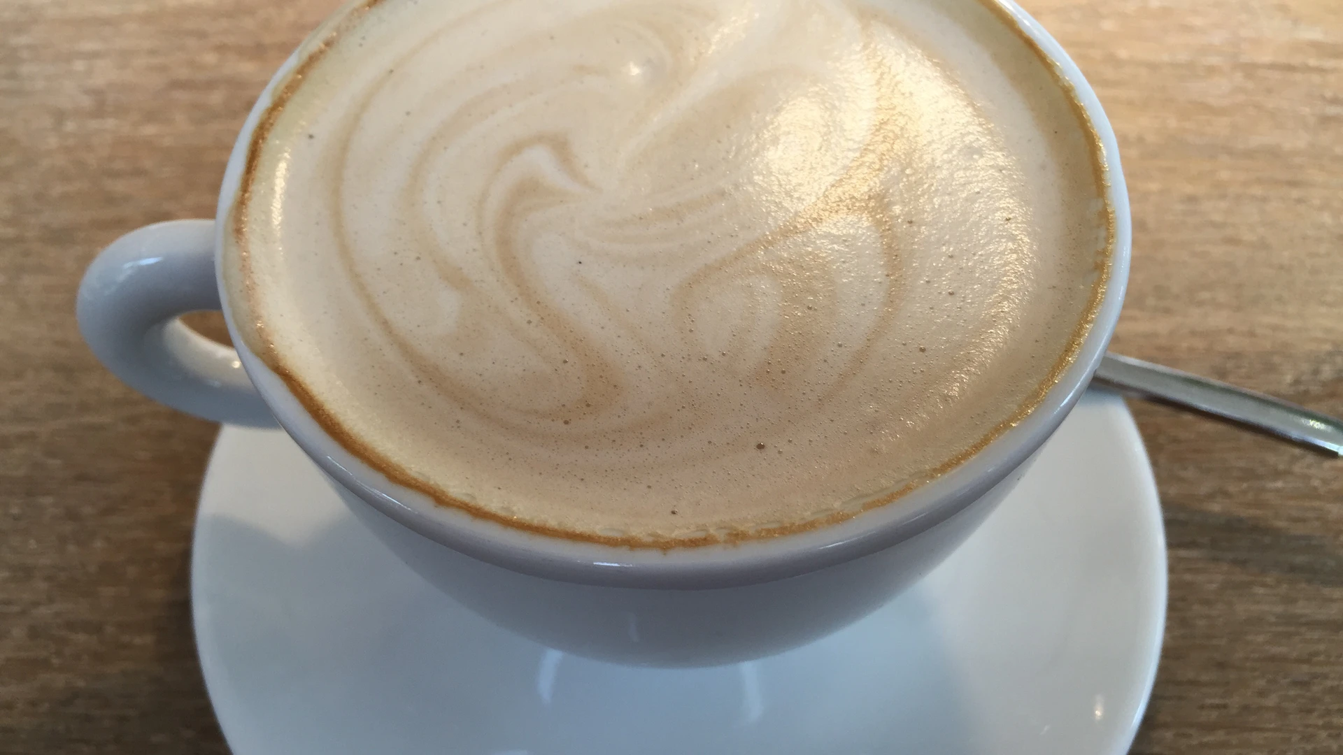 Coffee cup with frothy milk surface on a wooden table, accompanied by a teaspoon.