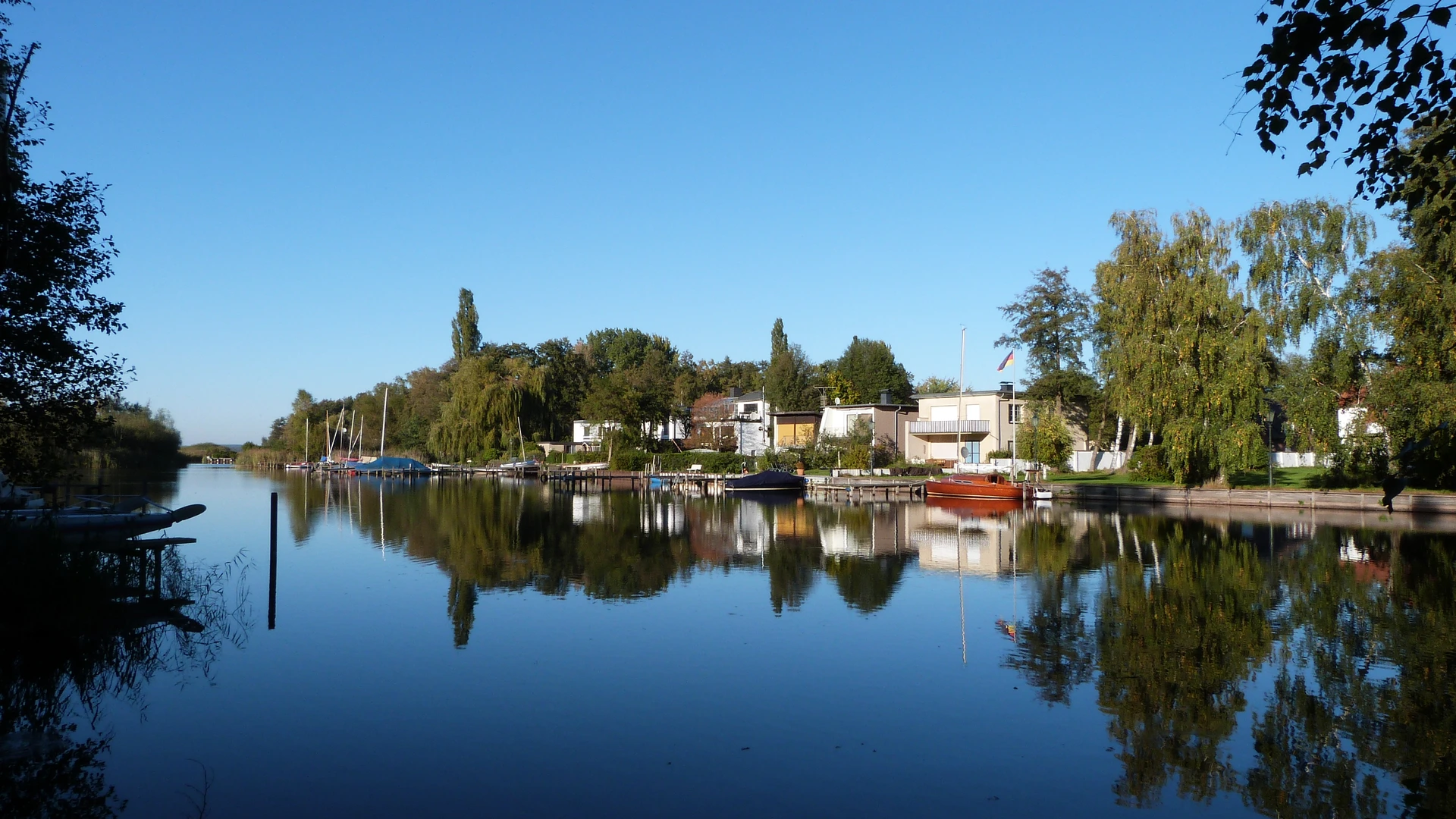 Gracht mit ruhigem Wasser, umgeben von Bäumen und Häusern im Grünen, unter klarem, blauem Himmel.
