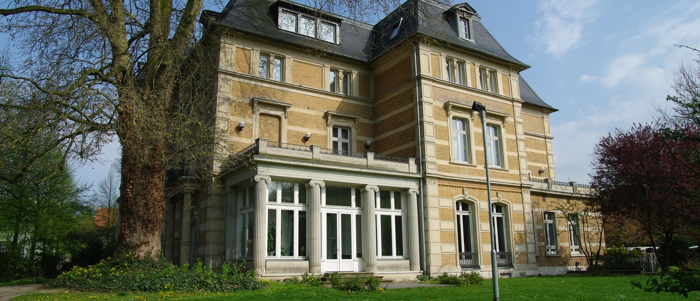 Villa Zanders Historic villa with a yellow facade, large windows and a mansard roof, surrounded by trees and lawn.