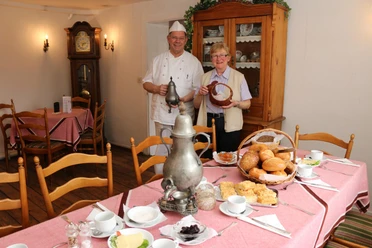 035 Krop. Berg. Kaffeetafel HN.JPG An elderly couple in a pub holds a bread basket; a table set with breakfast is in front of them.