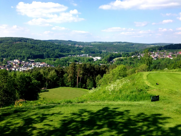 Bahn2__c__GC_Luederich.jfif Green golf course landscape in hilly surroundings with forest and village under blue sky, trees line the edge.
