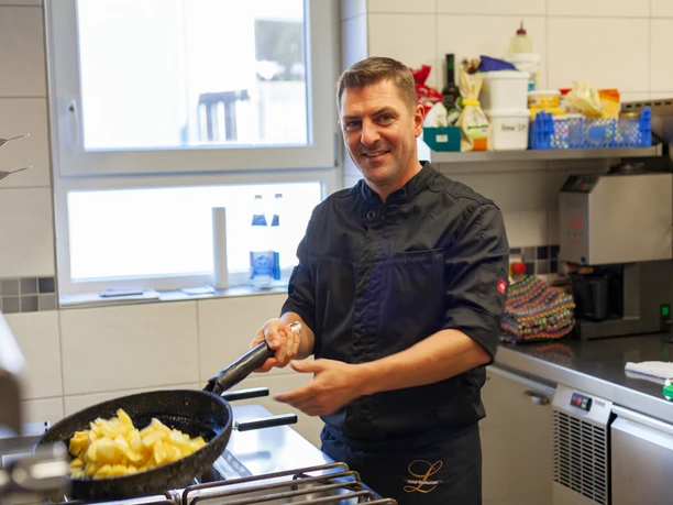 Hotel - Restaurant Lüdenbach A chef in a black uniform prepares a potato dish in a pan in a modern kitchen.
