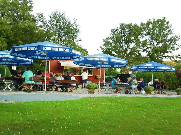 Hotel - Restaurant Lüdenbach A beer garden with blue umbrellas, people sitting at wooden tables, surrounded by green countryside.