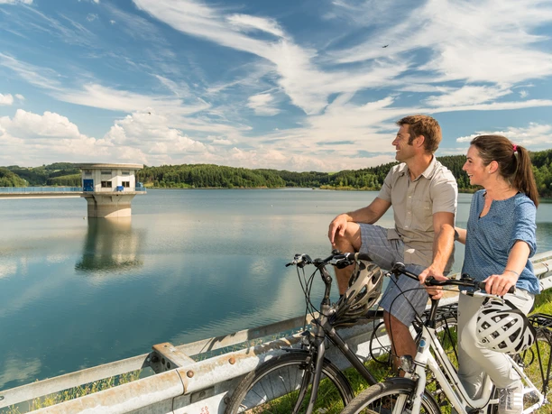 Cycling at the Dhünn Dam A couple on bicycles enjoys the view of a tranquil reservoir landscape on a sunny day.