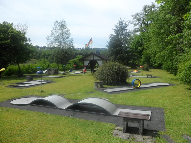 Minigolfbaan in Reichshof-Eckenhagen Minigolfbaan met een groene weide, bomen, kleurrijke hindernissen en een klein huisje op de achtergrond.