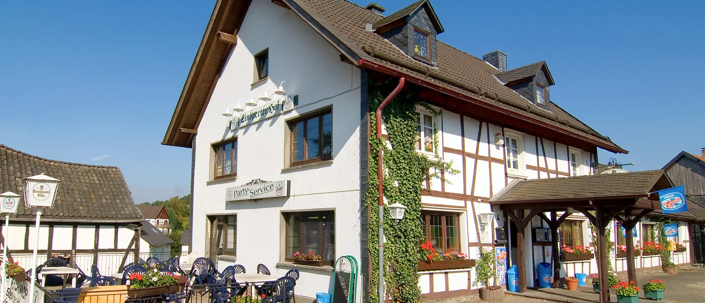 Sinsperter Hof A half-timbered house with a restaurant and a terrace full of plants under a clear, blue sky.