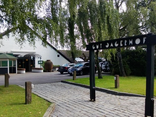 Steakhaus Gut Jägerhof w Erkrath Wejście do restauracji serwującej steki Gut Jägerhof w Erkrath z utwardzoną ścieżką, drzewami i zaparkowanymi samochodami.
