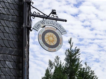 Zewnętrzny szyld Palazzo Wiedenhof w Haan Okrągły szyld Palazzo Wiedenhof w Haan z dekoracyjnym wzorem słońca, otoczony drzewami.