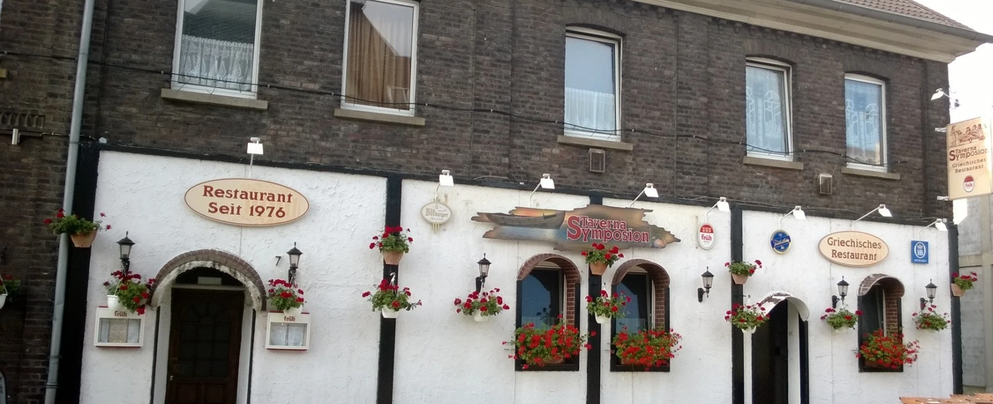 Sympozjum Tawerny Na zdjęciu widać tradycyjny budynek restauracji z białymi ścianami i ozdobnymi kwiatami.