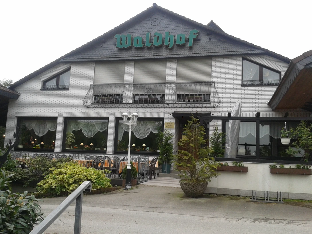 Kawiarnia Restauracja Waldhof Biały ceglany budynek z zielonym szyldem Waldhof, zasłoniętym oknem i latarnią na pierwszym planie.