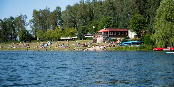 Sommertag am Badestrand des Stausee Oberwald mit Wiese, Badegästen und Bäumen im Hintergrund.