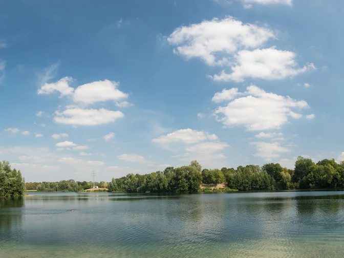 Lago Verde nel parco ricreativo Volkardey a Ratingen Un lago limpido circondato da rive fittamente boscose, con un cielo azzurro brillante sopra.